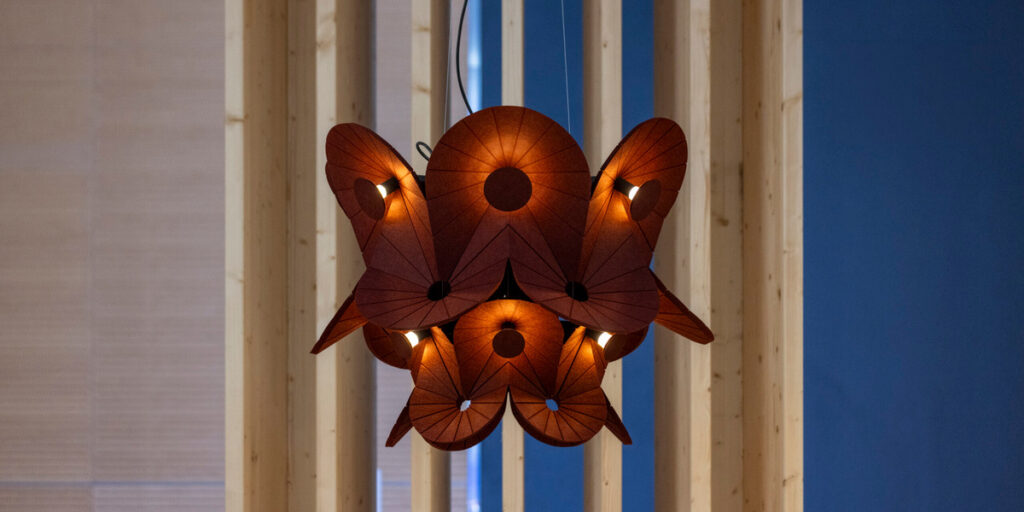 Luminaria colgante de tonos marrones con pantallas plegadas y luz cálida frente a una estructura de madera.
